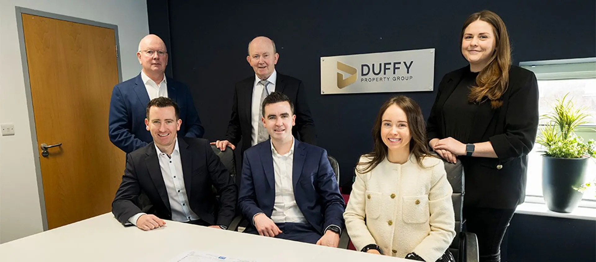 Six people in business attire pose around a white table in an office, with a DUFFY PROPERTY GROUP sign on the wall behind them. Three are seated, three stand, and plants line the windowsill—highlighting the team behind Duffy Capital’s success.