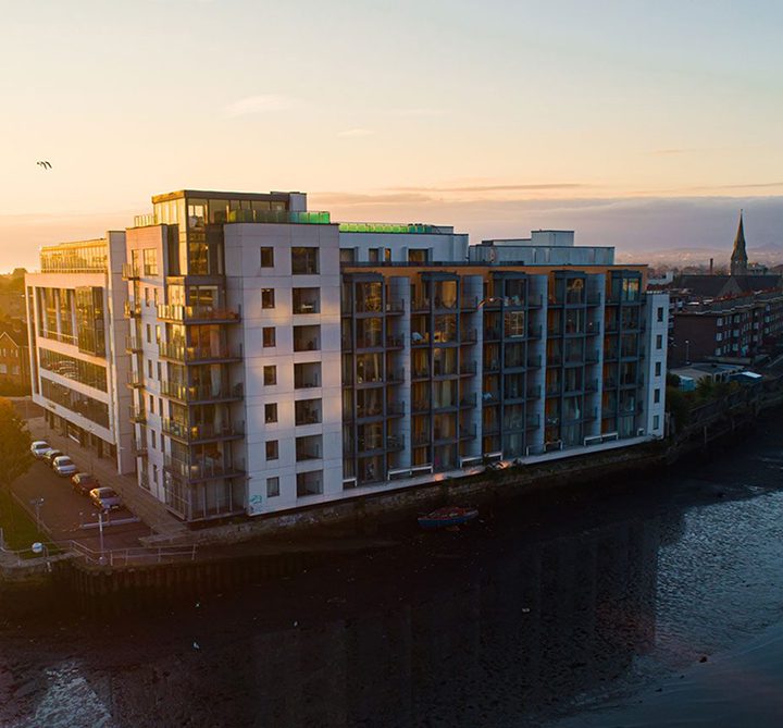 A modern multi-story apartment building by Duffy Capital, with large glass balconies aglow in the sunset, stands beside a body of water at low tide, with a distant town and church spire enhancing the scenic backdrop.
